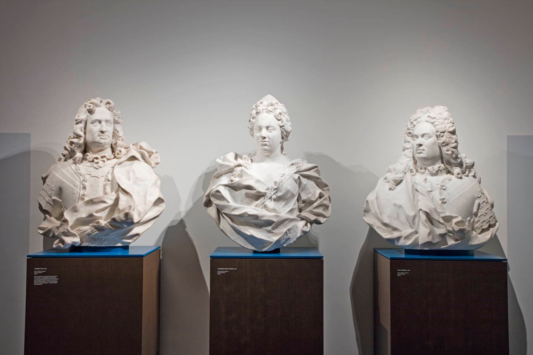 Drei Marmorbüsten historischer Persönlichkeiten mit aufwendiger Kleidung werden auf Sockeln in einem Museumsambiente ausgestellt.