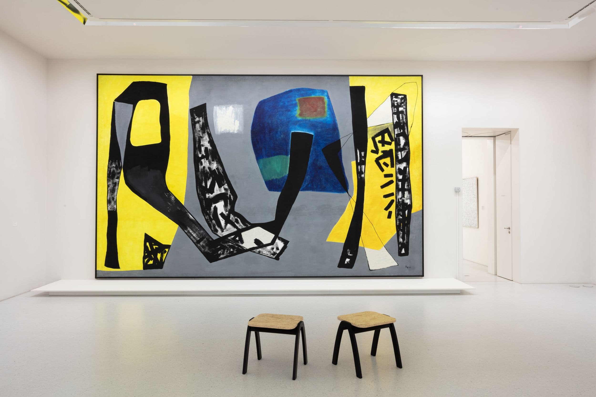 Abstraktes Gemälde mit kräftigen schwarzen, gelben und blauen Formen an einer Galeriewand. Zwei Hocker stehen davor auf einem hellgrauen Boden.