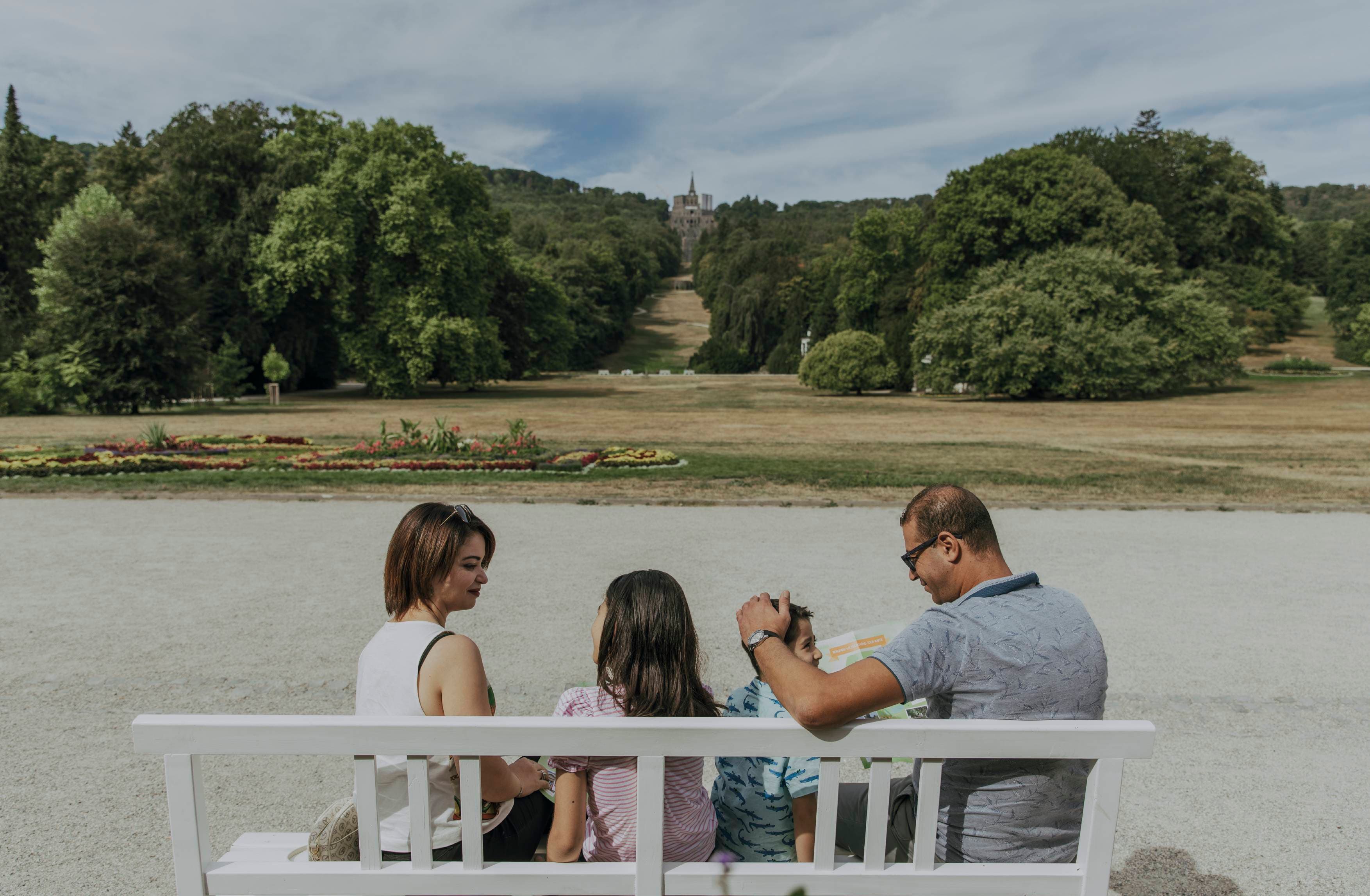 Vierköpfige Familie, die auf einer Bank im Bergpark sitzen; wobei sich im Hintergrund die Hauptachse des Bergparks bis zum Herkules erstreckt