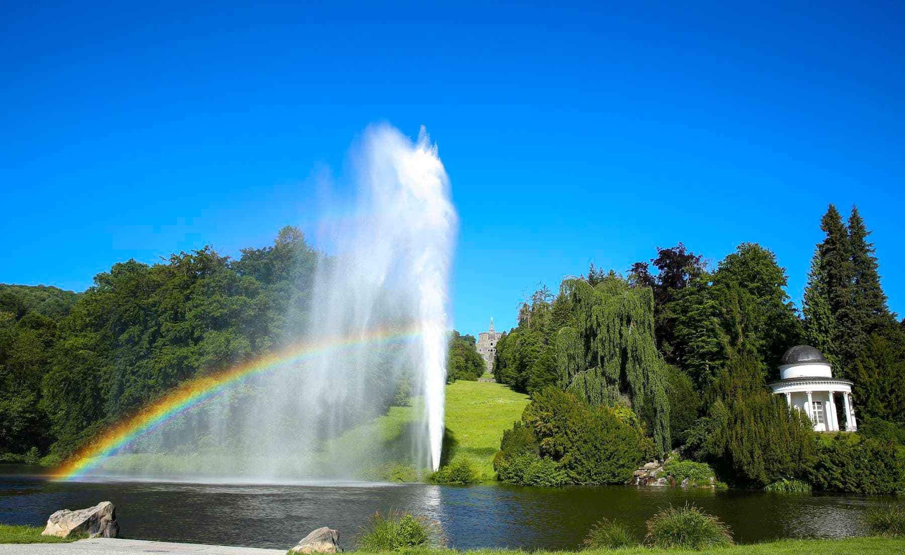 A vibrant rainbow arches over a large fountain in a lush park, with a white gazebo and dense trees under a clear blue sky.