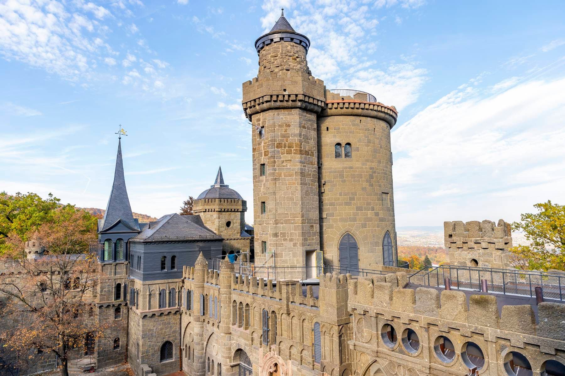 Historisches Schloss mit runden Türmen und zinnenbewehrten Mauern unter blauem Himmel, umgeben von herbstlichen Bäumen.