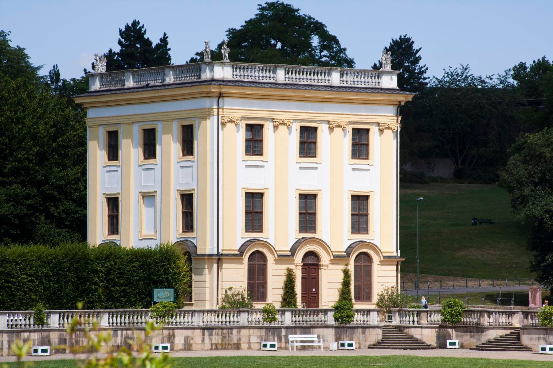 Ein gelb-weißes neoklassizistisches Gebäude mit Bogenfenstern und Flachdach, umgeben von Grün und einer Steinterrasse.