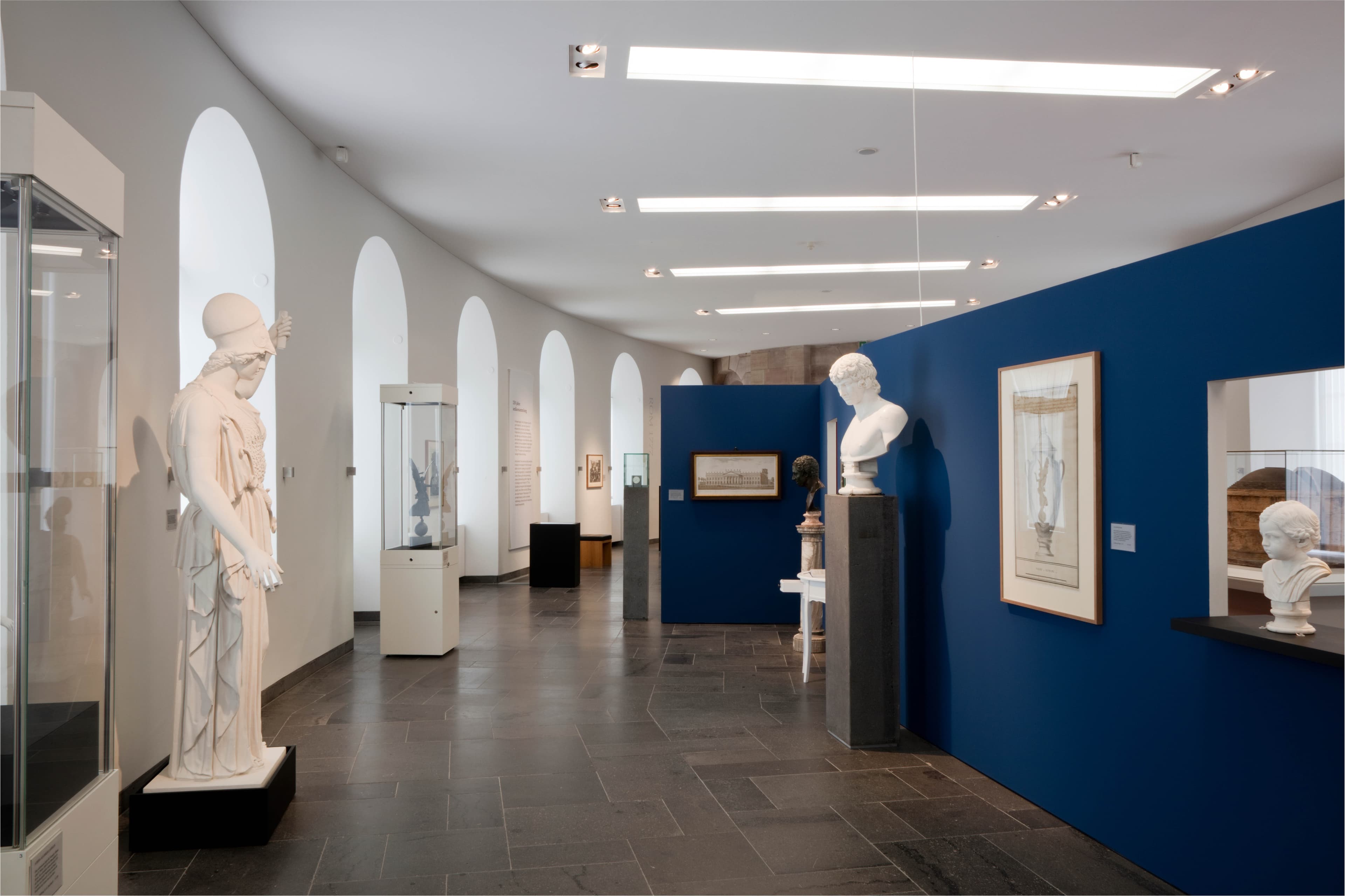 Museumsgalerie mit weißen Skulpturen, blauen Wänden und gerahmten Kunstwerken. Bogenfenster auf der linken Seite erhellen den Raum.